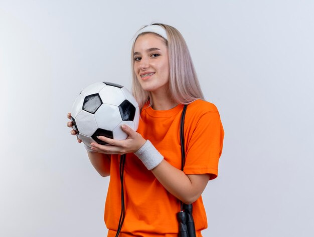 Sonriente joven caucásica deportiva con tirantes y con saltar la cuerda alrededor del cuello con diadema y muñequeras sostiene la bola en blanco