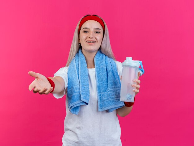 Sonriente joven caucásica deportiva con tirantes vistiendo muñequeras de diadema y con una toalla alrededor del cuello sostiene una botella de agua estirando la mano