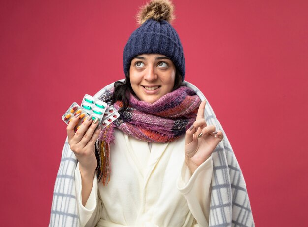 Sonriente joven caucásica enferma con bata de invierno sombrero y bufanda envuelta en cuadros sosteniendo paquetes de cápsulas mirando y apuntando hacia arriba aislado en la pared carmesí