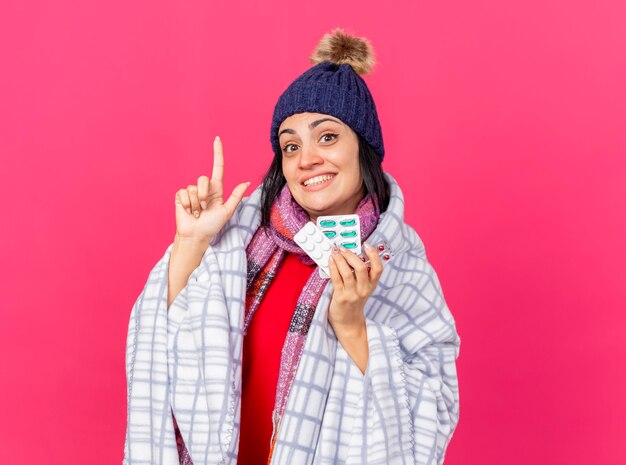 Sonriente joven caucásica enferma con gorro de invierno y bufanda envuelta en cuadros sosteniendo paquetes de píldoras médicas apuntando hacia arriba aislado en la pared carmesí con espacio de copia