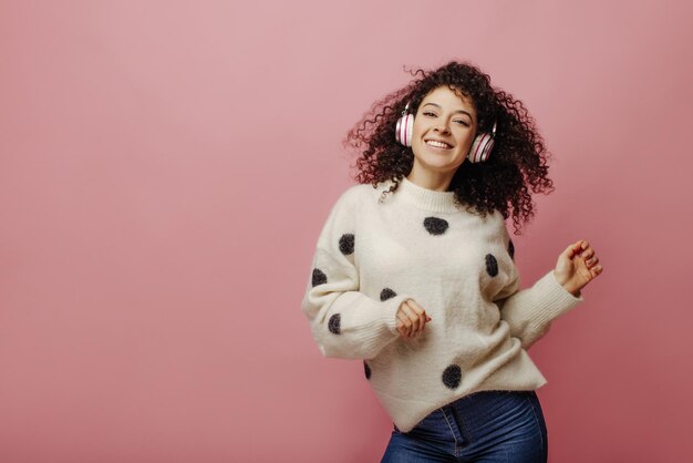 Sonriente joven caucásica está disfrutando de su canción favorita y bailando sobre fondo rosa Retrato de mujer morena rizada divirtiéndose en auriculares Tecnología de audio y concepto de música