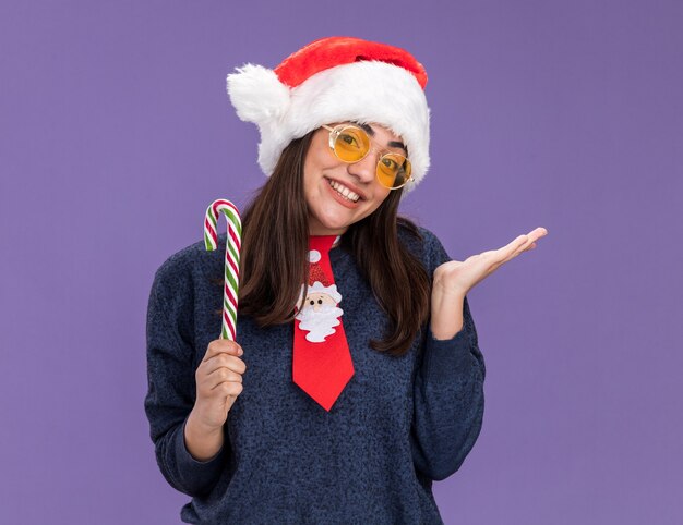 Sonriente joven caucásica en gafas de sol con gorro de Papá Noel y corbata de santa sostiene bastón de caramelo y mantiene la mano abierta aislada sobre fondo púrpura con espacio de copia