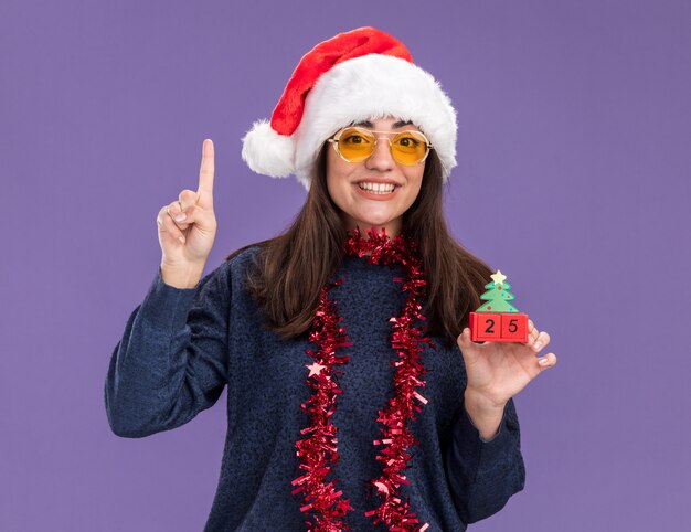 Sonriente joven caucásica en gafas de sol con gorro de Papá Noel y guirnalda alrededor del cuello sostiene adornos de árbol de Navidad y apunta hacia arriba aislado en la pared púrpura con espacio de copia