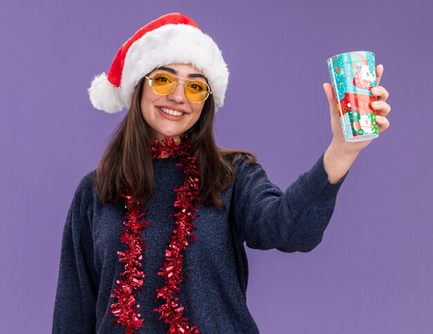 Sonriente joven caucásica en gafas de sol con gorro de Papá Noel y guirnalda alrededor del cuello sostiene un vaso de papel aislado en la pared púrpura con espacio de copia