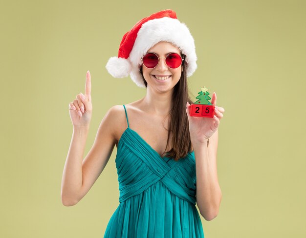 Sonriente joven caucásica en gafas de sol con gorro de Papá Noel sosteniendo el adorno del árbol de Navidad y apuntando hacia arriba