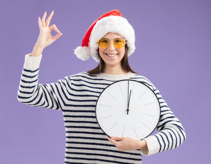 Foto gratuito sonriente joven caucásica en gafas de sol con sombrero de santa sosteniendo el reloj y gesticulando signo ok