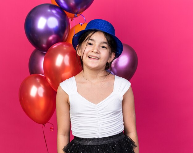 Sonriente joven caucásica con gorro de fiesta azul de pie delante de globos de helio aislado en la pared rosa con espacio de copia