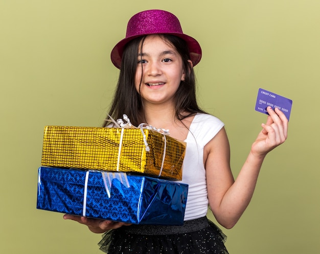 Sonriente joven caucásica con gorro de fiesta púrpura sosteniendo cajas de regalo y tarjeta de crédito aislado en la pared verde oliva con espacio de copia