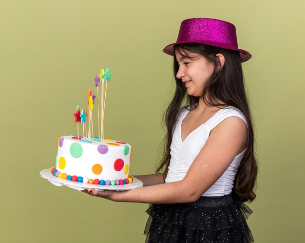 Sonriente joven caucásica con gorro de fiesta púrpura sosteniendo y mirando el pastel de cumpleaños de pie lateralmente aislado en la pared verde oliva con espacio de copia