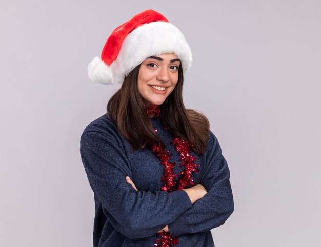 Sonriente joven caucásica con gorro de Papá Noel y guirnalda alrededor del cuello se encuentra con los brazos cruzados