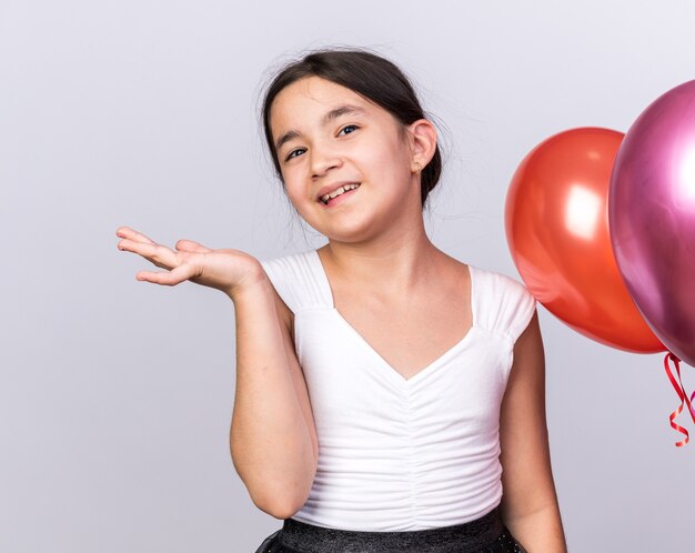 Sonriente joven caucásica de pie con globos de helio manteniendo la mano abierta aislada en la pared blanca con espacio de copia