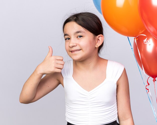 Sonriente joven caucásica de pie con globos de helio pulgar hacia arriba aislado en la pared blanca con espacio de copia