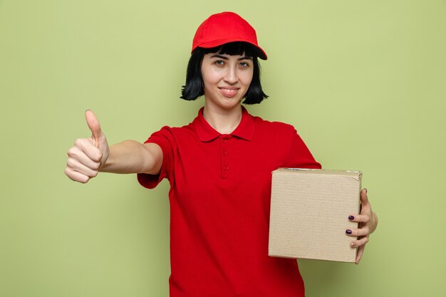 Sonriente joven caucásica repartidora sosteniendo una caja de cartón y pulgar hacia arriba