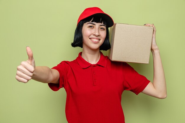 Sonriente joven caucásica repartidora sosteniendo una caja de cartón sobre su hombro y apuntando hacia arriba