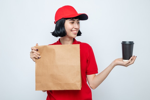 Sonriente joven caucásica repartidora sosteniendo envases de alimentos y mirando la taza de papel