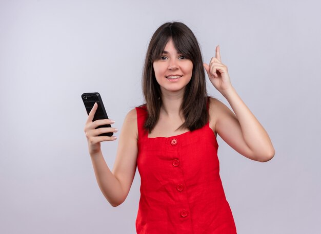 Sonriente joven caucásica sosteniendo teléfono apunta con el dedo hacia arriba y mirando a cámara sobre fondo blanco aislado