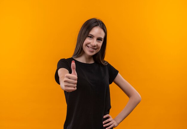 Sonriente joven caucásica vistiendo camiseta negra con el pulgar hacia arriba puso la mano en la cadera sobre fondo naranja aislado