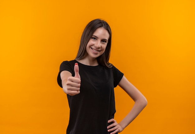 Sonriente joven caucásica vistiendo camiseta negra con el pulgar hacia arriba puso la mano en la cadera sobre fondo naranja aislado