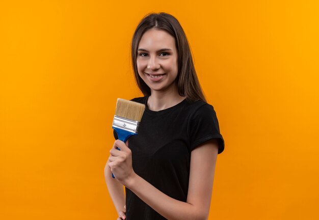 Sonriente joven caucásica vistiendo camiseta negra sosteniendo pincel sobre fondo naranja aislado