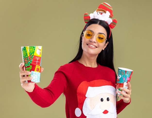 Sonriente joven caucásica vistiendo santa claus diadema y suéter con gafas sosteniendo vasos plásticos de Navidad estirando uno mirando aislado sobre fondo verde oliva