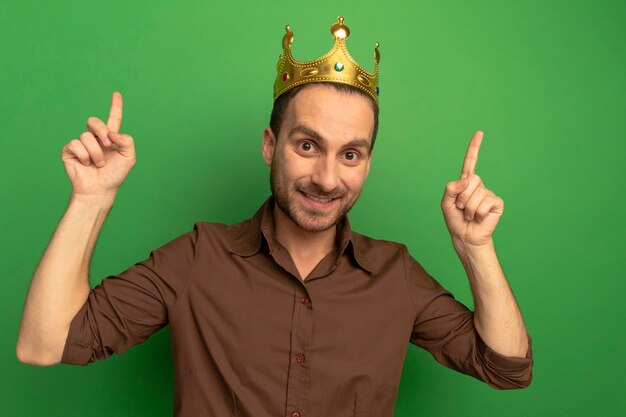 Sonriente joven caucásico con corona apuntando hacia arriba aislado en la pared verde