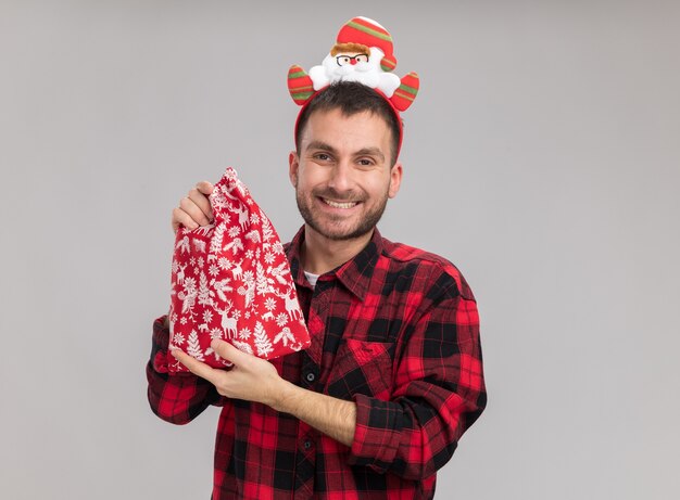 Sonriente joven caucásico con diadema de Navidad sosteniendo saco de Navidad mirando a cámara aislada sobre fondo blanco con espacio de copia