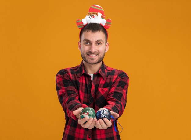 Sonriente joven caucásico con diadema de santa claus mirando a la cámara estirando adornos navideños hacia la cámara aislada sobre fondo naranja con espacio de copia