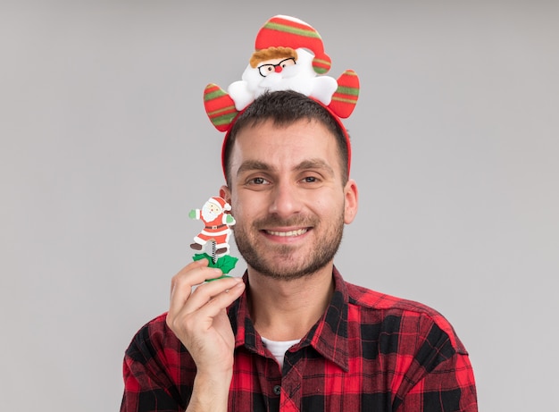 Sonriente joven caucásico con diadema de santa claus sosteniendo muñeco de nieve juguete de navidad mirando a cámara aislada sobre fondo blanco