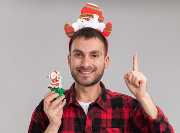 Sonriente joven caucásico con diadema de santa claus sosteniendo muñeco de nieve juguete de navidad mirando a la cámara apuntando hacia arriba aislado sobre fondo blanco.