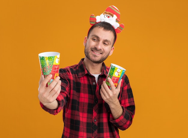 Sonriente joven caucásico con diadema de santa claus sosteniendo vasos de Navidad de plástico estirando uno mirando aislado sobre fondo naranja con espacio de copia