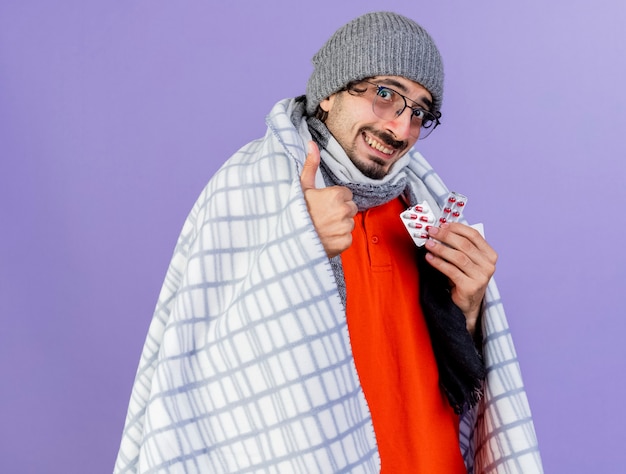 Sonriente joven caucásico enfermo con gafas, gorro de invierno y bufanda envuelto en cuadros sosteniendo paquetes de cápsulas mirando a cámara mostrando el pulgar hacia arriba aislado sobre fondo púrpura