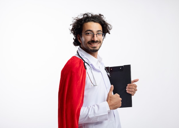 Sonriente joven caucásico con gafas ópticas con uniforme médico con manto rojo y con estetoscopio alrededor del cuello se encuentra de lado sosteniendo el portapapeles y los pulgares hacia arriba