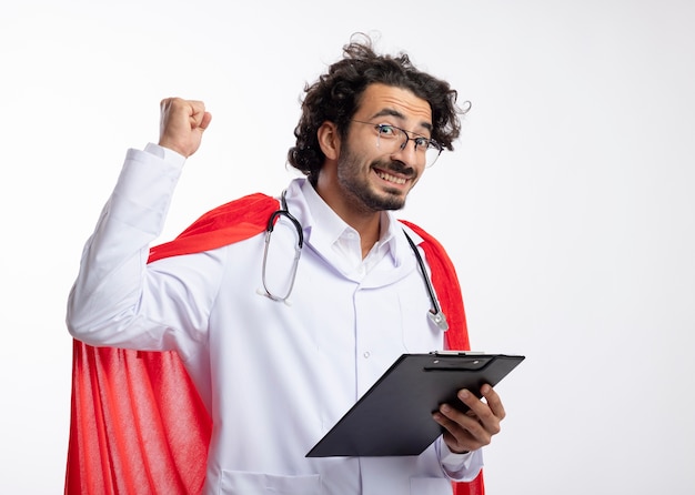 Sonriente joven caucásico en gafas ópticas con uniforme médico con manto rojo y con estetoscopio alrededor del cuello se encuentra con el puño levantado y sostiene el portapapeles en la pared blanca