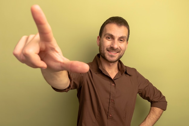 Sonriente joven caucásico manteniendo la mano en la cintura mirando a la cámara estirando la mano haciendo gesto de perdedor aislado sobre fondo verde oliva