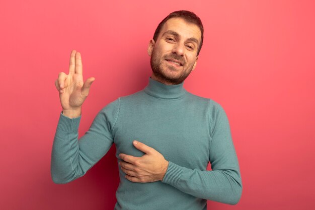 Sonriente joven caucásico mirando a la cámara haciendo gesto de pistola manteniendo la mano en el pecho aislado sobre fondo carmesí con espacio de copia