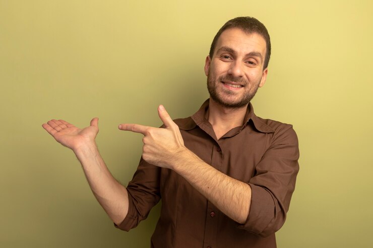 Foto gratuito sonriente joven caucásico mirando a la cámara mostrando la mano vacía apuntando aislado sobre fondo verde oliva