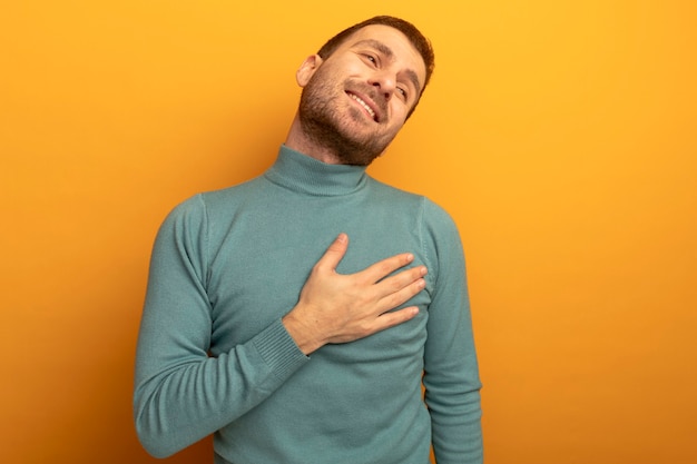 Sonriente joven caucásico poniendo la mano en el pecho mirando al lado aislado en la pared naranja con espacio de copia