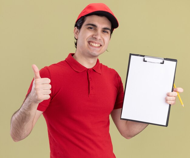 sonriente, joven, caucásico, repartidor, en, uniforme rojo, y, gorra, actuación, portapapeles, mostrando, pulgar, arriba, con, lápiz, en, otro, mano, aislado, en, verde oliva, pared