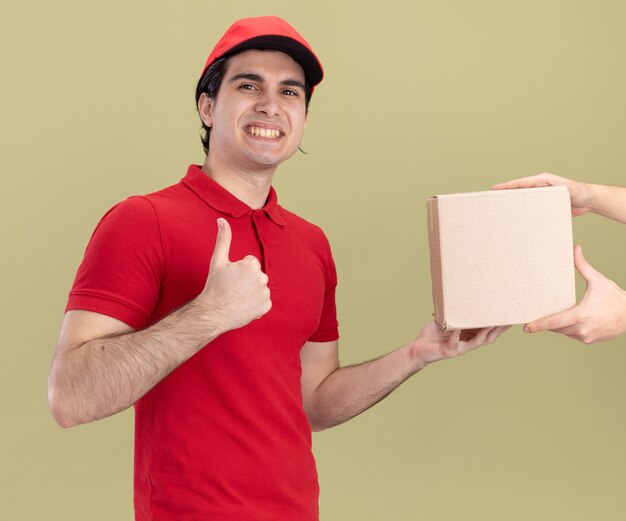 Sonriente joven caucásico repartidor en uniforme rojo y gorra dando cardbox al cliente mostrando el pulgar hacia arriba aislado en la pared verde oliva