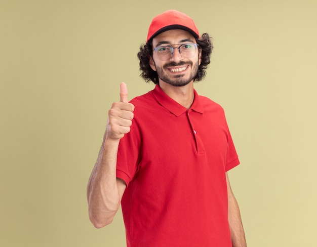 Sonriente joven caucásico repartidor en uniforme rojo y gorra con gafas mostrando el pulgar hacia arriba aislado en la pared verde oliva con espacio de copia