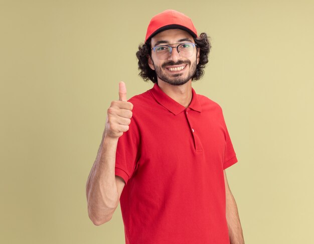 Sonriente joven caucásico repartidor en uniforme rojo y gorra con gafas mostrando el pulgar hacia arriba aislado en la pared verde oliva con espacio de copia
