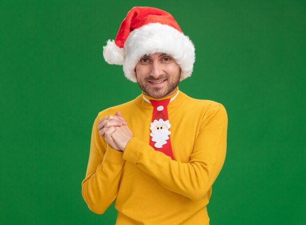 Sonriente joven caucásico con sombrero de navidad y corbata mirando a la cámara haciendo gesto ganador aislado sobre fondo verde con espacio de copia
