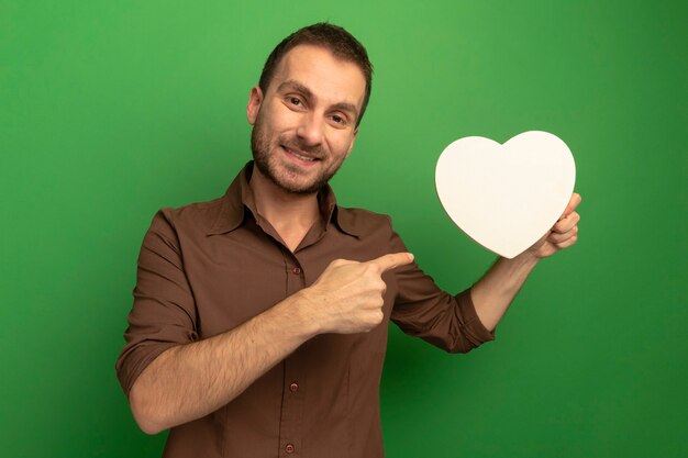 Sonriente joven caucásico sosteniendo y apuntando a la forma del corazón mirando a la cámara aislada sobre fondo verde