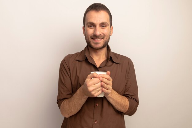 Sonriente joven caucásico sosteniendo una taza de té mirando a la cámara aislada sobre fondo blanco con espacio de copia