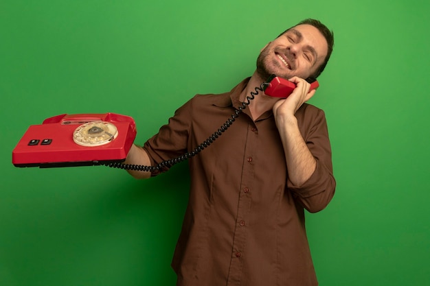 Sonriente joven caucásico sosteniendo teléfono antiguo hablando por teléfono con los ojos cerrados aislado sobre fondo verde con espacio de copia