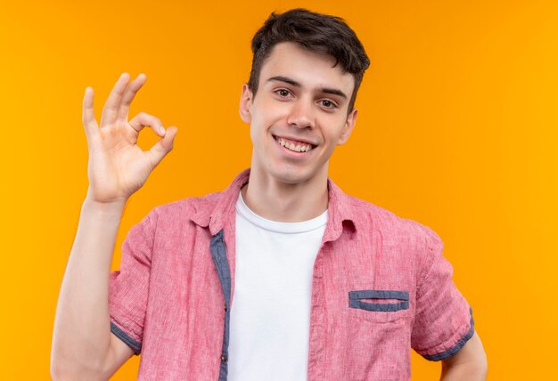 Sonriente joven caucásico vistiendo camisa rosa mostrando gesto okey sobre fondo naranja aislado