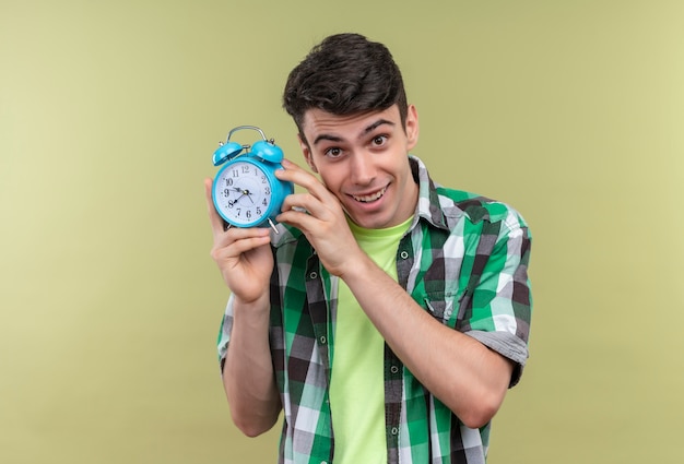 Sonriente joven caucásico vistiendo camisa verde sosteniendo el reloj de alarma alrededor de la cara con ambas manos sobre fondo verde aislado