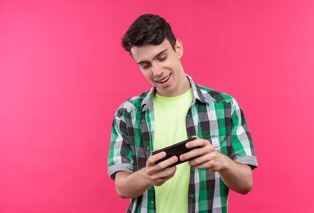 Sonriente joven caucásico vistiendo la camiseta verde jugando en el teléfono sobre fondo rosa aislado