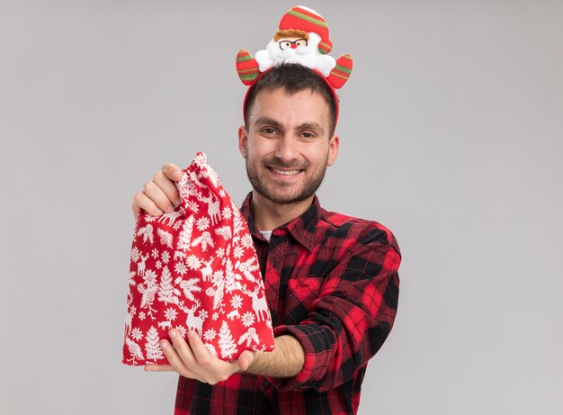 Sonriente joven caucásico vistiendo diadema navideña estirando el saco de navidad hacia la cámara mirando a cámara aislada sobre fondo blanco con espacio de copia