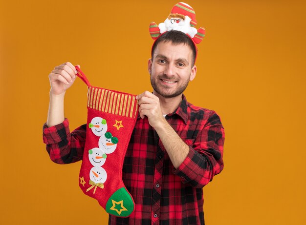 Sonriente joven caucásico vistiendo santa claus diadema sosteniendo calcetines de Navidad mirando a cámara aislada sobre fondo naranja con espacio de copia
